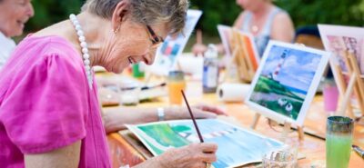 Happy, smiling elderly woman painting a landscape during an art class, illustrating the mentally stimulating activities and social maintenance provided by assisted living for the elderly.