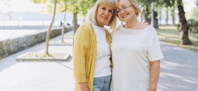 Two smiling senior women walking arm-in-arm in a park, representing companionship for seniors and quality elder care services.